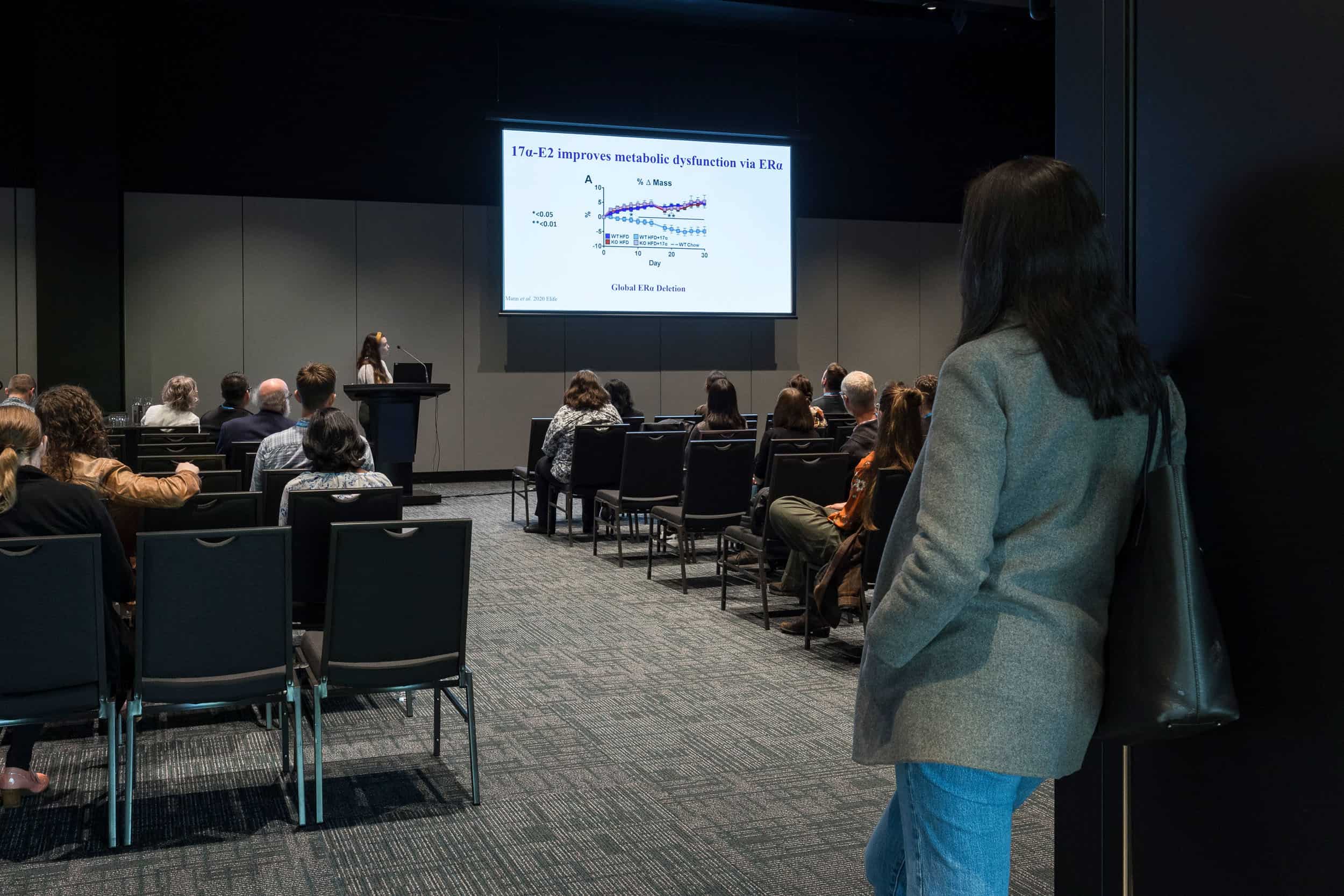 Ateendee leans on doorway watching speaker during medical conference presentation, Te Pae, Christchurch.