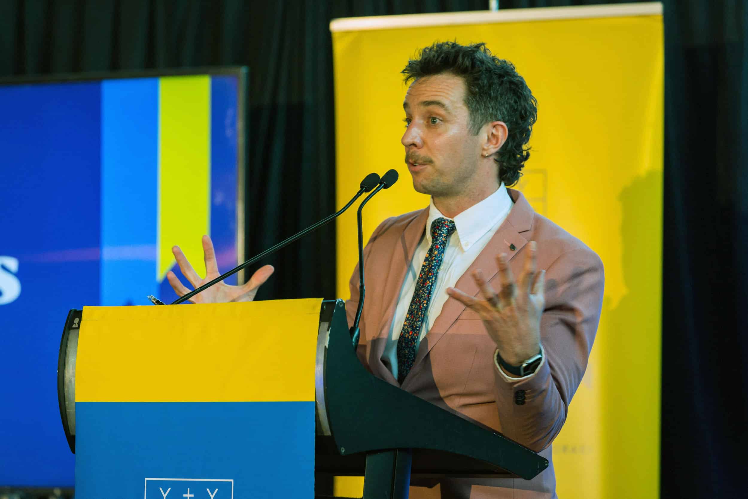 Rhys Mathewson speaks from a podium as the MC at a dinner event.