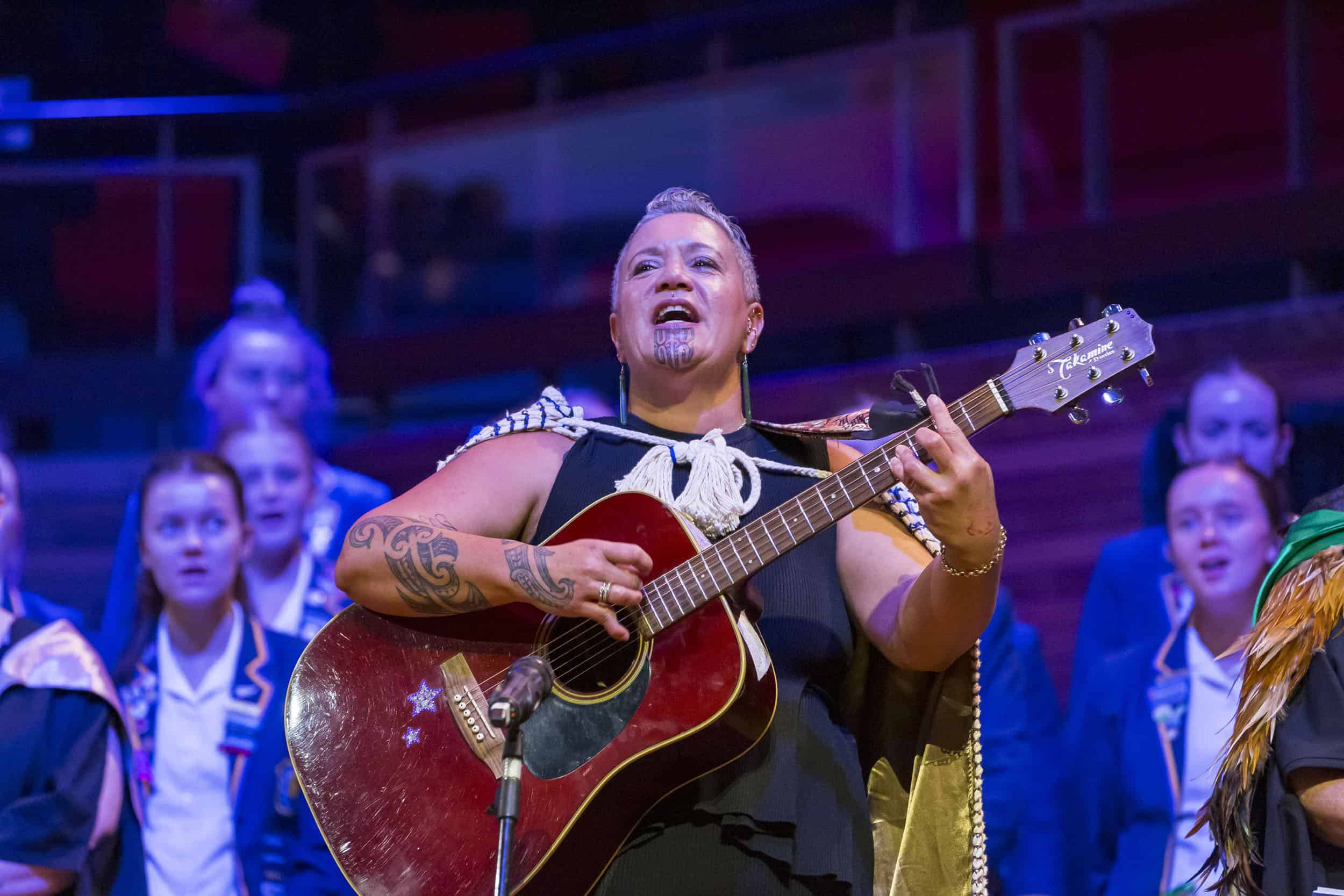 Maori teacher with moko leads a waiata playing guitar at a school prizegiving Christchurch Town Hall.