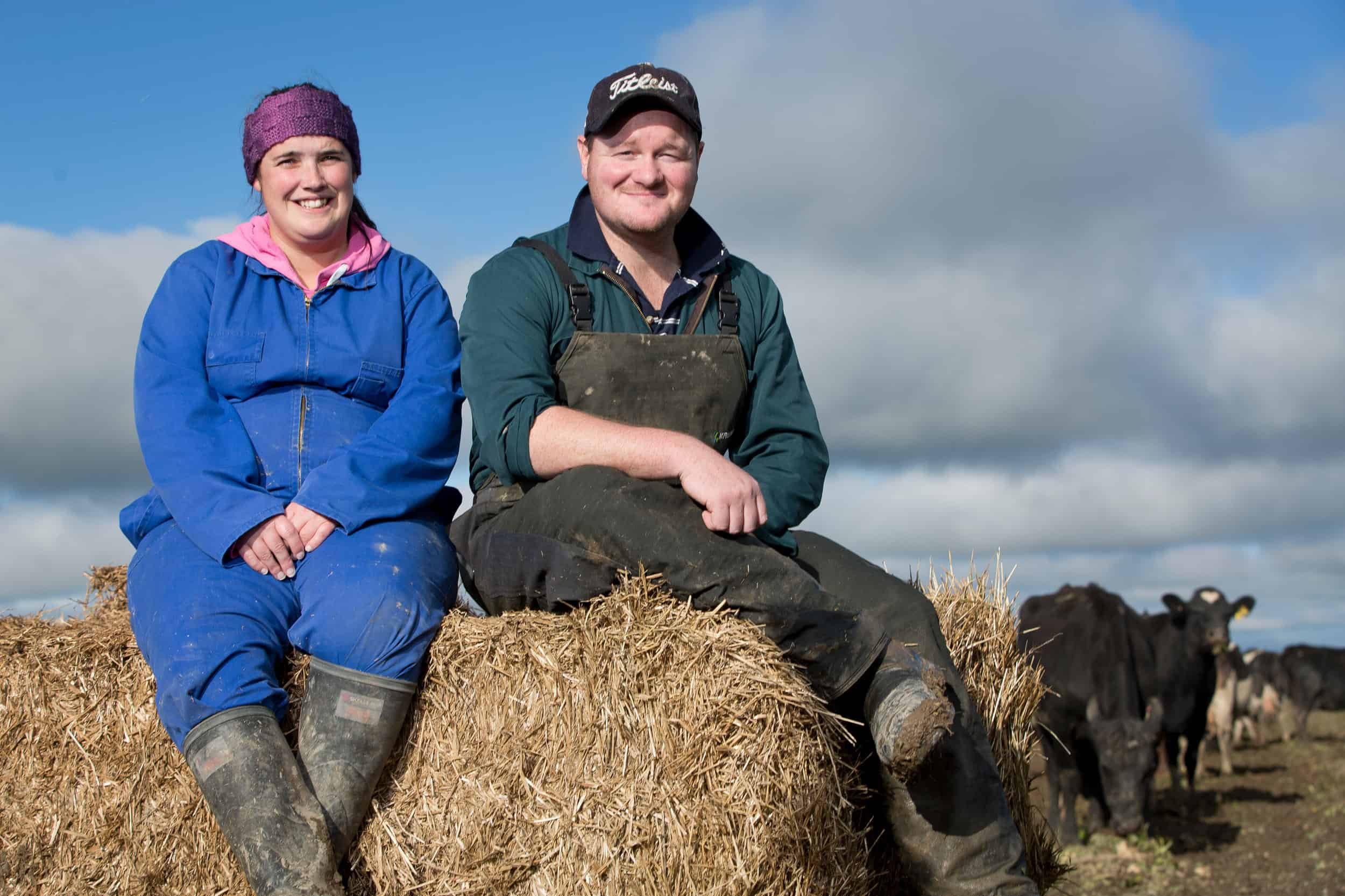 Dairy farming couple sit on haybales with cows behind.