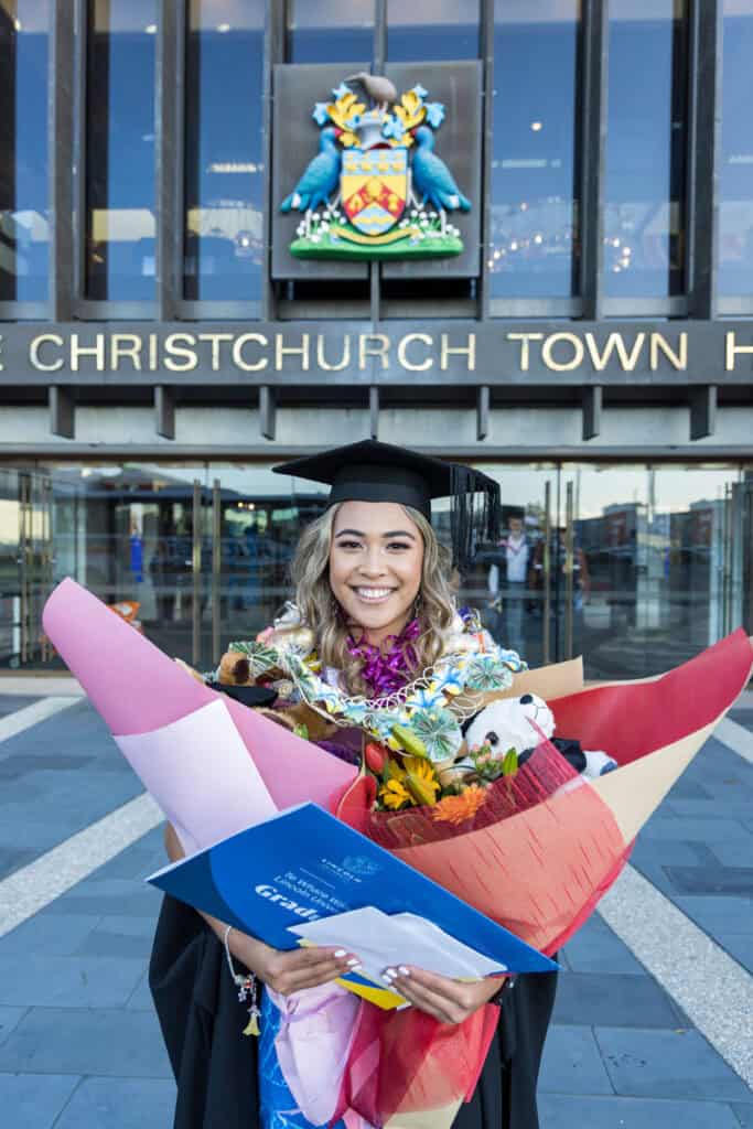 Young female graduate adorned with bouquets poses outside the CHCH Town Hall.